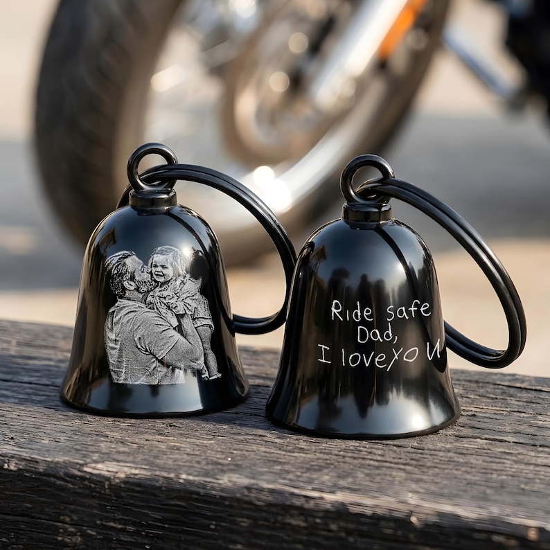 May include: Two black motorcycle bells with a loop for hanging. One bell features an engraved image of a man holding a child. The other bell has the text "Ride safe Dad, I love you".