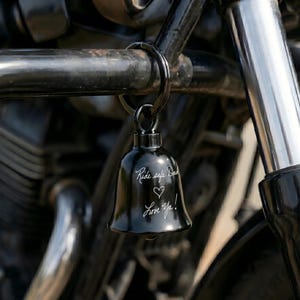 May include: A black motorcycle bell with the inscription "Ride safe Dad" and a heart symbol, followed by "Love Ya!" The bell is suspended from a black metal ring, attached to a motorcycle. The bell is a small, decorative item.