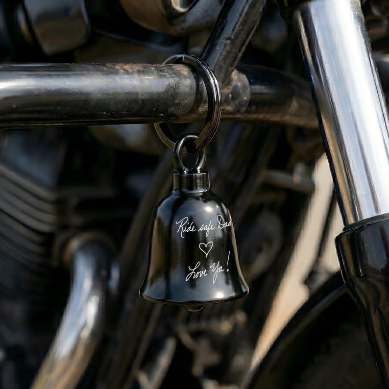 May include: A black motorcycle bell with the inscription "Ride safe Dad" and a heart symbol, followed by "Love Ya!" The bell is suspended from a black metal ring, attached to a motorcycle. The bell is a small, decorative item.