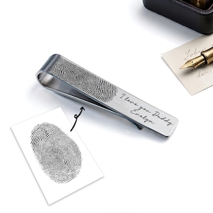May include: A silver-colored tie clip with a fingerprint design and the engraved text "I love you Daddy Evelyn." A small card with a fingerprint is next to the tie clip. A pen and paper are in the background.