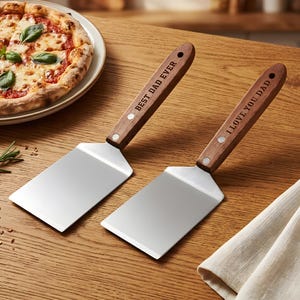 May include: Two stainless steel pizza spatulas with wooden handles. One spatula reads "BEST DAD EVER" and the other reads "I LOVE YOU DAD". The spatulas are on a wooden surface near a pizza.