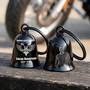 May include: Two black motorcycle bells with a loop for hanging. One bell has a motorcycle with wings and the text "RIDING WITH ANGELS" and "Lucas Ruverson 1978-2026". The other bell has a portrait of a man.