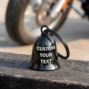 May include: A black motorcycle bell with the text "CUSTOM YOUR TEXT" in white. The bell is on a wooden surface, with a motorcycle wheel in the background. The bell has a loop for hanging.