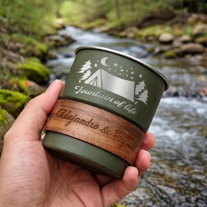 K&ouml;nnte beinhalten: Ein olivgr&uuml;ner Campingbecher mit silbernem Rand und Holzmanschette. Der Becher zeigt eine Campingszene mit B&auml;umen, einem Zelt und dem Text "Fountain of life". Die Holzmanschette ist mit "Alejandro & Papi" graviert.