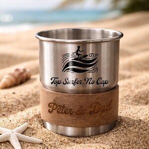 K&ouml;nnte beinhalten: Ein Edelstahlbecher mit einer schwarzen Grafik eines Surfers und dem Text "Top Surfer, No Cap". Der Boden ist mit einem braunen Material mit dem eingravierten Text "Peter & Dad" umwickelt. Der Becher steht am Sandstrand.