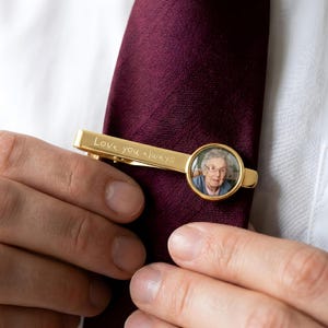 May include: A gold-colored tie clip with the engraved text "Love you always." The clip features a circular photo of a person. The tie clip is attached to a burgundy tie.