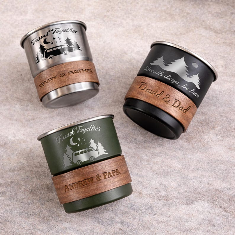 K&ouml;nnte beinhalten: Drei Reisebecher mit eingravierten Designs. Einer ist silberfarben, einer schwarz und einer olivgr&uuml;n. Jeder Becher hat ein Holzband mit personalisiertem Text. Der silberne Becher tr&auml;gt die Aufschrift "Travel Together" und "HENRY & FATHER". Der schwarze Becher tr&auml;gt die Aufschrift "Breath deep, be here" und "David & Dad". Der olivgr&uuml;ne Becher tr&auml;gt die Aufschrift "Travel Together" und "ANDREW & PAPA".