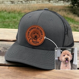May include: Gray trucker hat with a black mesh back and a circular leather patch. The patch features a dog portrait and the text "THIS HUMAN BELONGS TO Charles". The hat is on a wooden surface.