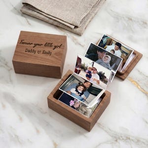May include: A wooden photo box with the inscription "Forever your little girl Daddy Emily" on the lid. The box is open, revealing a series of photographs. A folded linen cloth rests in the background.