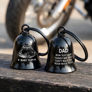May include: Two black motorcycle bells with a loop for hanging. One bell has a baby ultrasound image and the text "BABY TOM". The other bell says "DAD Now you can carry me too can't wait to ride with you".
