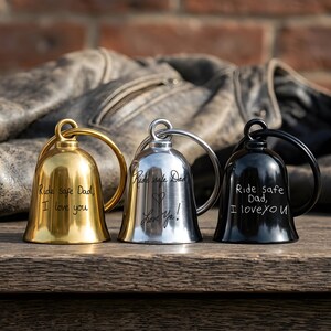 May include: Three motorcycle bells in gold, silver, and black. Each bell has a loop for hanging and features the text "Ride safe Dad, I love you" or similar sentiments. The bells are displayed on a wooden surface.