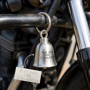 May include: A silver motorcycle bell with the inscription "Ride safe Dad, I love you" is attached to a motorcycle. The bell is suspended from a metal ring. A small card with the same message is attached.