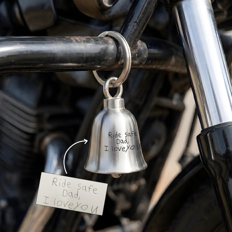 May include: A silver motorcycle bell with the inscription "Ride safe Dad, I love you" is attached to a motorcycle. The bell is suspended from a metal ring. A small card with the same message is attached.