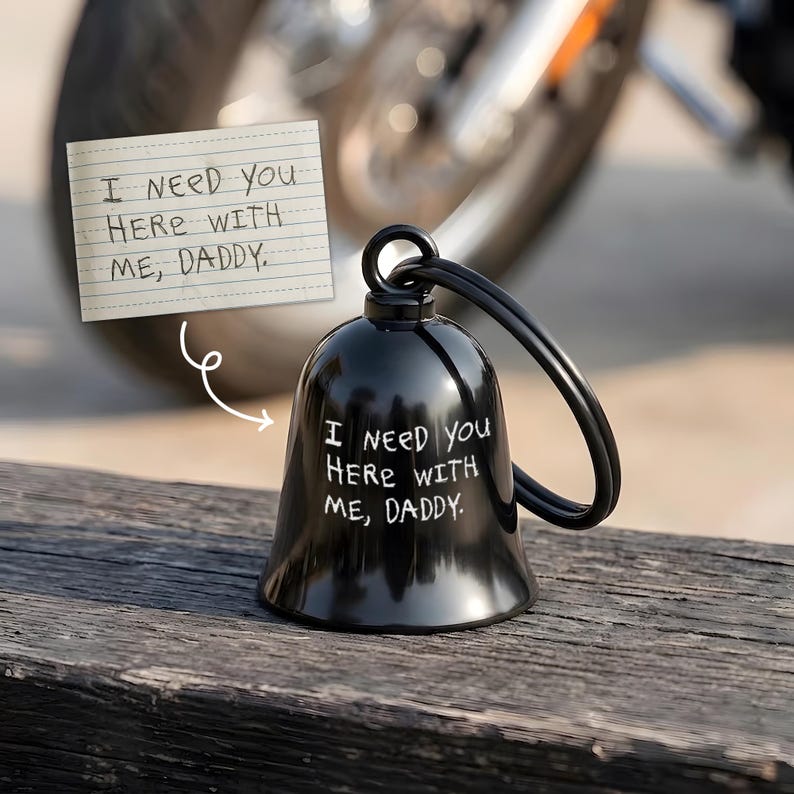 May include: Black bell-shaped motorcycle bell with a black handle. The bell has the text "I NEED YOU HERE WITH ME, DADDY" in white. A small notepad with the same text is in the background.