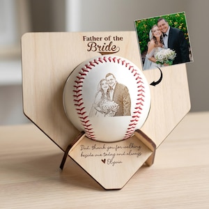 May include: A baseball with a laser-etched wedding photo, displayed on a wooden home plate-shaped stand. The baseball has red stitching and the words "Father of the Bride" are above it. The stand has a photo and a message.
