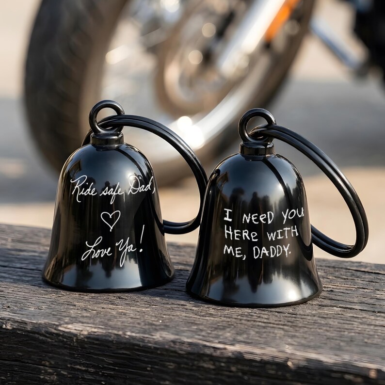 May include: Two black motorcycle bells with white handwritten text. One bell reads "Ride safe Dad, Love Ya!" with a heart symbol. The other bell says "I need you here with me, Daddy."