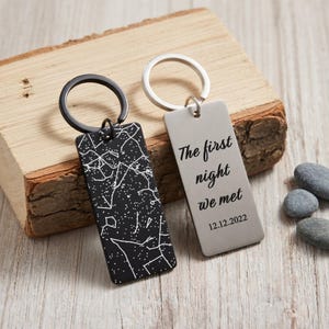 May include: Two rectangular keychains. One is black with a white constellation design. The other is silver with the text "The first night we met" and the date 12.12.2022. Both have metal key rings.