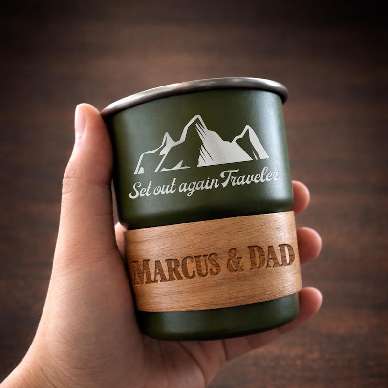 K&ouml;nnte beinhalten: Ein olivgr&uuml;ner Reisebecher mit silbernem Rand und Holzband. Der Becher zeigt eine wei&szlig;e Berg-Grafik und den Text "Set out again Traveler" und "MARCUS & DAD".