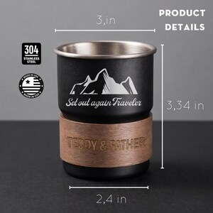 K&ouml;nnte beinhalten: Schwarzer und silberner Reisebecher mit Holzsockel. Der Becher zeigt eine Berg-Grafik und den Text "Set out again Traveler". Er ist 8,48 cm hoch, 7,62 cm breit und hat einen Durchmesser von 6,1 cm. Der Sockel ist mit "TEDDY & FATHER" graviert.