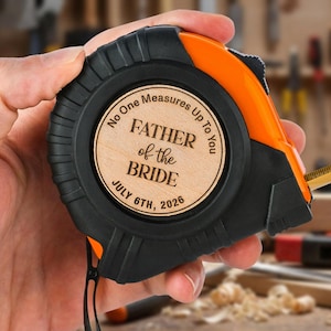 May include: A black and orange tape measure featuring a circular wooden inlay engraved with the text No One Measures Up To You, FATHER of the BRIDE, and the date JULY 6TH, 2026, centered on the side of the tool casing.
