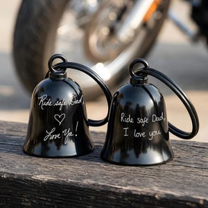 May include: Two black motorcycle bells with the text "Ride safe Dad" and "Love Ya!" and "Ride safe Dad, I love you" in white script. The bells have a loop for hanging and are displayed on a wooden surface.