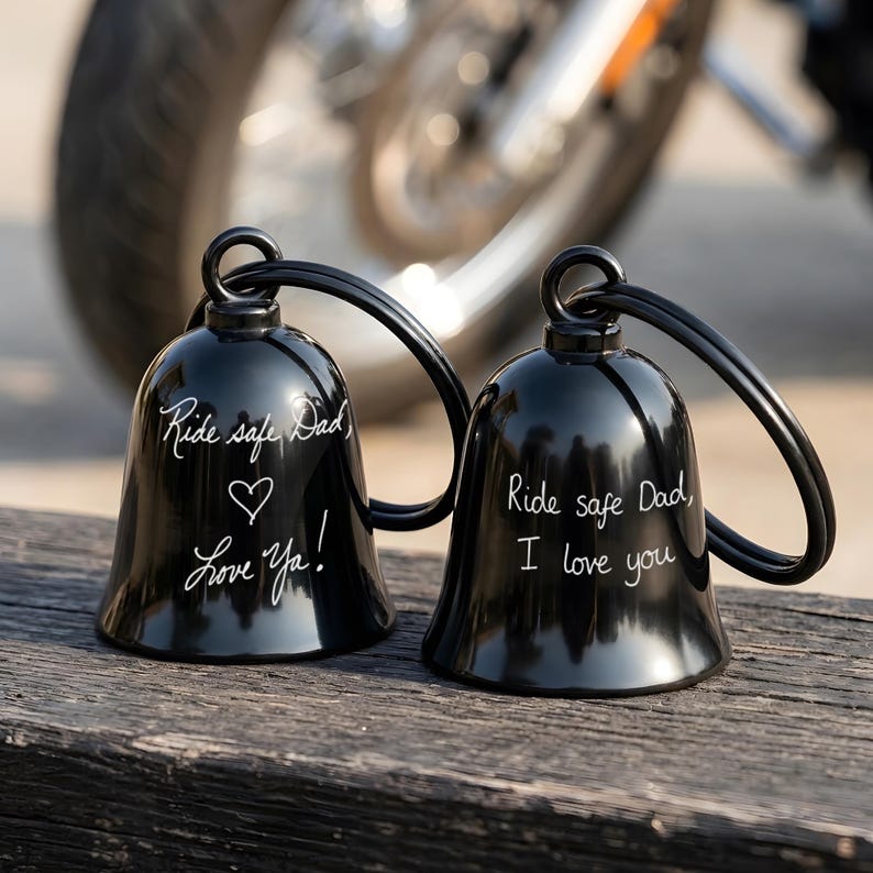 May include: Two black motorcycle bells with the text "Ride safe Dad" and "Love Ya!" and "Ride safe Dad, I love you" in white script. The bells have a loop for hanging and are displayed on a wooden surface.