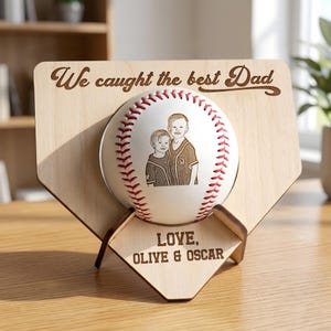 May include: A light-colored wooden home plate display stand featuring a white baseball with red stitching. The wood is engraved with the text "We caught the best Dad" and "LOVE, OLIVE & OSCAR". The baseball features a brown line-art illustration.