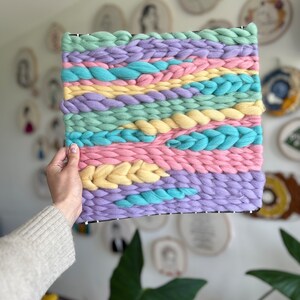 May include: A square woven wall hanging with a pastel color scheme. The piece features horizontal bands of light blue, pink, yellow, and lavender yarn, with braided and textured details. The yarn is woven onto a black frame.