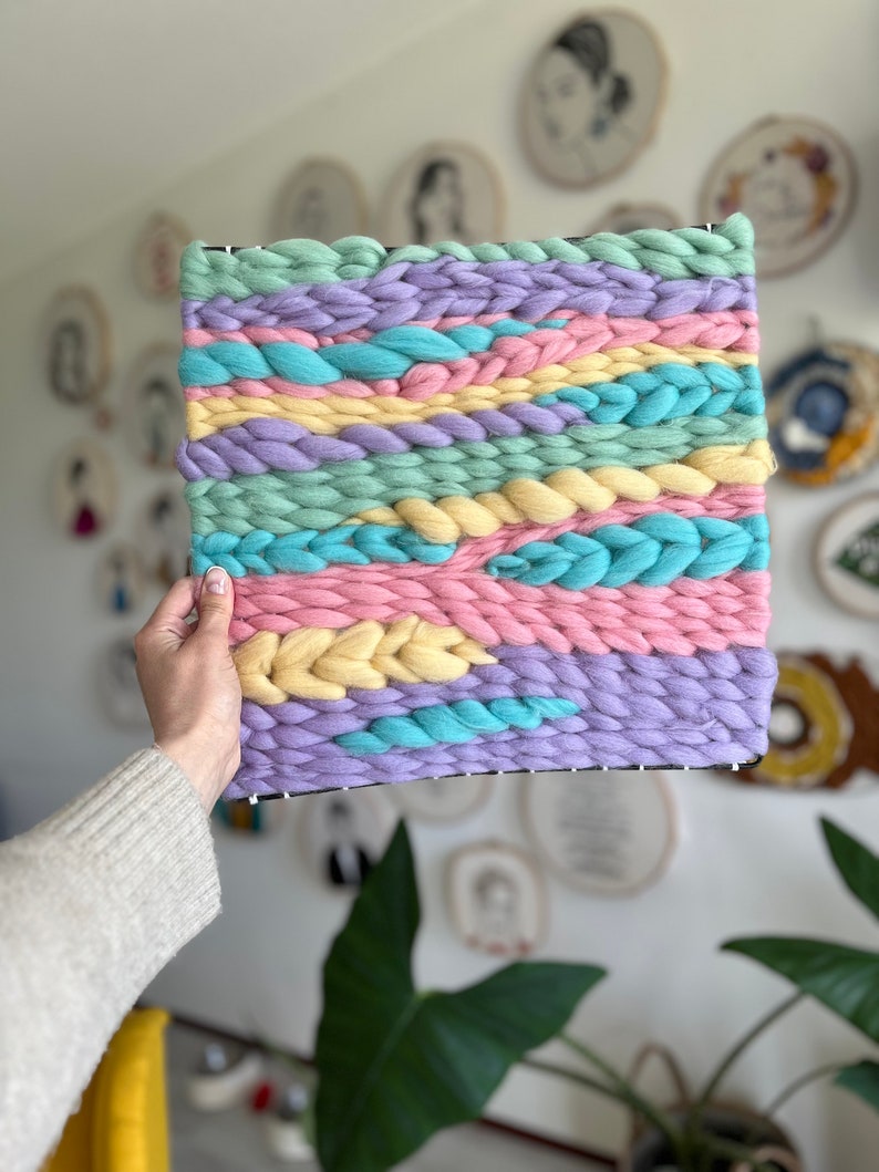 May include: A square woven wall hanging with a pastel color scheme. The piece features horizontal bands of light blue, pink, yellow, and lavender yarn, with braided and textured details. The yarn is woven onto a black frame.