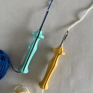 Puede incluir: Dos herramientas de aguja de hilo con mangos azul y amarillo. La aguja azul tiene un hilo azul enhebrado y la aguja amarilla tiene un hilo blanco enhebrado. Una bola de hilo azul está a la izquierda de la aguja azul y una bola de hilo blanco está a la derecha de la aguja amarilla.