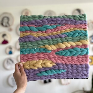 May include: A square woven wall hanging with a black frame. The textile art piece features horizontal bands of pastel colors, including light green, lavender, pink, yellow, and turquoise. The woven texture is thick and chunky, creating a tactile and visually appealing design.