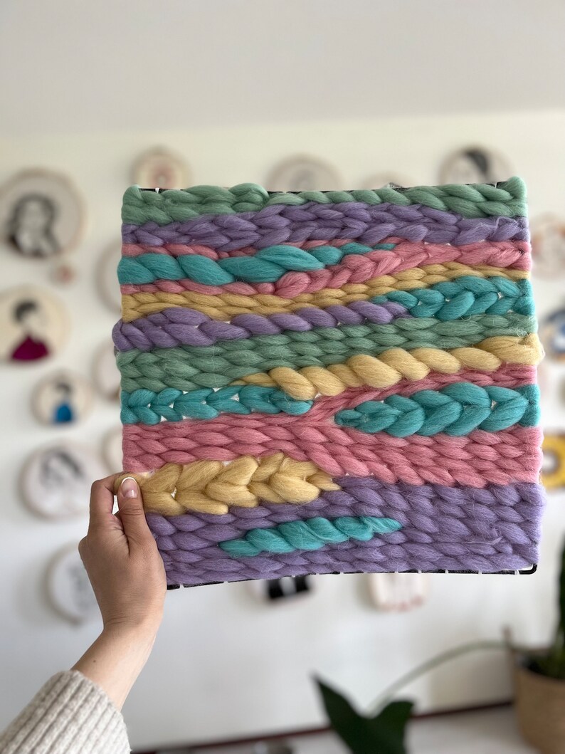 May include: A square woven wall hanging with a black frame. The textile art piece features horizontal bands of pastel colors, including light green, lavender, pink, yellow, and turquoise. The woven texture is thick and chunky, creating a tactile and visually appealing design.