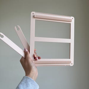 Op de afbeelding: Een lichtroze weefgetouw met twee bijpassende spoelen. Het weefgetouw heeft een rechthoekig frame met gelijkmatig verdeelde tanden langs de boven- en onderranden. De spoelen zijn plat en peddelvormig. Ambachtelijke benodigdheden.