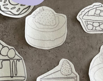 Op de afbeelding: Geborduurde strijk-op stickers met verschillende dessertontwerpen, waaronder een cupcake, donut, aardbeientaart, taart en twee verschillende taartpunten. De stickers zijn wit met zwarte borduurwerk.