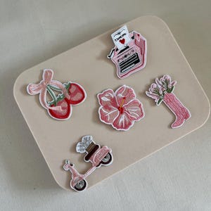 Puede incluir: Seis parches de hierro bordados de color rosa. Los parches incluyen una máquina de escribir, una cereza, una flor de hibisco, un ramo de flores, un scooter y un corazón con el texto "Te amo".