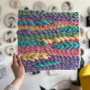 May include: A square, woven wall hanging with a black frame. The textile art piece features horizontal bands of pastel colors, including light green, pink, yellow, blue, and purple. The woven texture is thick and chunky, creating a tactile and visually appealing design.