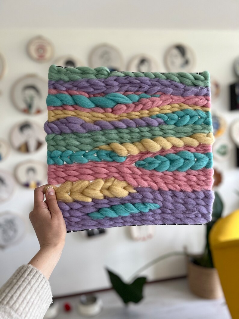 May include: A square, woven wall hanging with a black frame. The textile art piece features horizontal bands of pastel colors, including light green, pink, yellow, blue, and purple. The woven texture is thick and chunky, creating a tactile and visually appealing design.