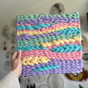 May include: A woven wall hanging with a rectangular shape. The piece features horizontal bands of pastel colors, including light blue, pink, yellow, green, and purple. The woven texture is visible, with thick yarn creating a tactile surface. The piece is held by a hand.