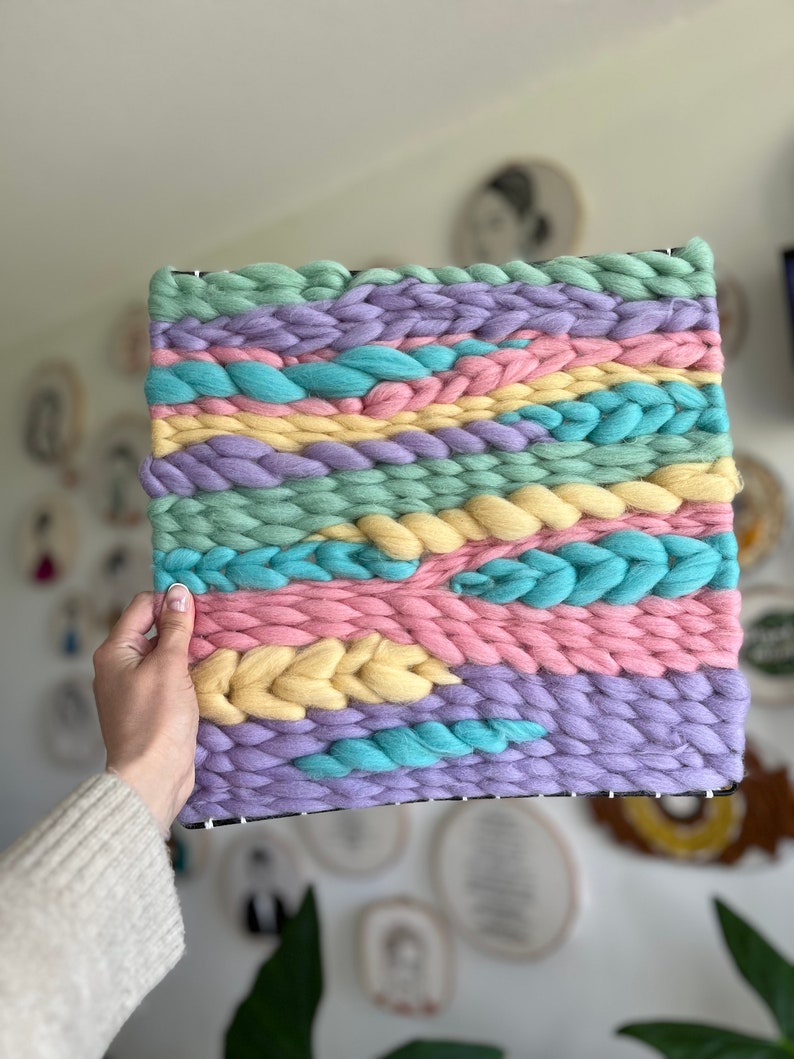 May include: A woven wall hanging with a rectangular shape. The piece features horizontal bands of pastel colors, including light blue, pink, yellow, green, and purple. The woven texture is visible, with thick yarn creating a tactile surface. The piece is held by a hand.