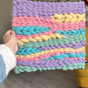 May include: A colorful woven wall hanging in pastel shades of lavender, pink, yellow, and teal. The chunky yarn is woven in horizontal bands, creating a textured, abstract design. The piece is rectangular and mounted on a black frame.