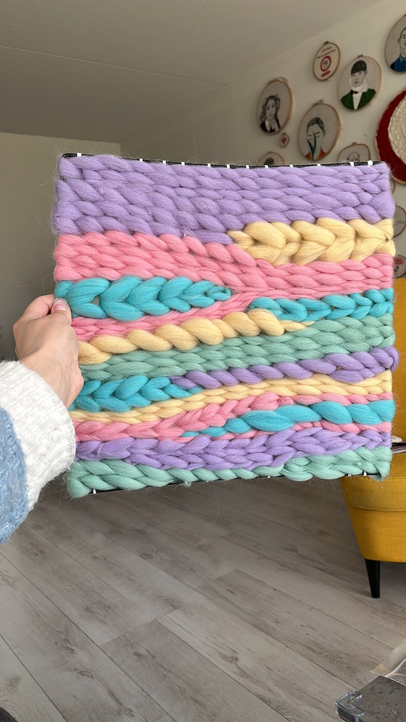 May include: A colorful woven wall hanging in pastel shades of lavender, pink, yellow, and teal. The chunky yarn is woven in horizontal bands, creating a textured, abstract design. The piece is rectangular and mounted on a black frame.
