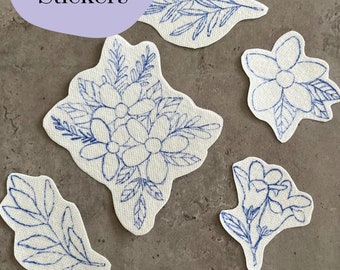 Könnte beinhalten: Fünf weiße Stickaufkleber mit blauen Nähten. Die Aufkleber zeigen florale Designs, darunter Blätter und Blumen.