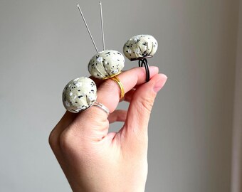 Può includere: Tre cuscinetti per spilli in tessuto con motivi floreali sono attaccati ad anelli, indossati su un dito. I cuscinetti per spilli sono beige con fiori bianchi e accenti neri. Due dei cuscinetti per spilli hanno aghi inseriti.