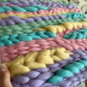 May include: A close-up of a handwoven textile in pastel colors. The piece features thick, braided strands of yarn in shades of lavender, pink, yellow, teal, and green. The texture is chunky and the overall effect is soft and inviting.