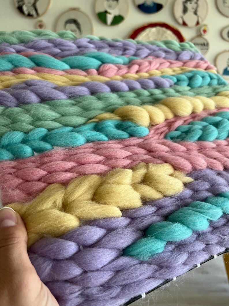 May include: A close-up of a handwoven textile in pastel colors. The piece features thick, braided strands of yarn in shades of lavender, pink, yellow, teal, and green. The texture is chunky and the overall effect is soft and inviting.