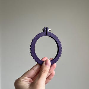 Op de afbeelding: Een paarse borduurring met een geschulpte rand. De ring wordt in een hand gehouden en heeft een metalen sluiting om stof vast te zetten. Het ronde frame heeft een diameter van ongeveer 10 cm.