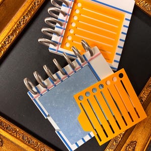 May include: Two spiral-bound notebooks with white covers featuring blue and pink stripes. One notebook has an orange checklist insert, while the other features a blue rectangular insert and a separate orange stencil with circular cutouts.