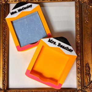 May include: Two pencil-shaped sticky note holders with orange bodies, pink tips, and white tops featuring black text that reads Mrs. McCauley and Mrs. Beacher. The holders have a square cutout for paper and are displayed within a gold ornate frame.