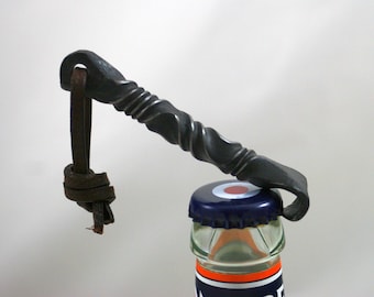 May include: A black metal bottle opener with a twisted handle and a leather loop. The opener is being used to open a bottle of beer.