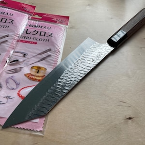 May include: A Japanese-style kitchen knife with a hammered metal blade and a dark wooden handle, positioned next to two clear plastic packages containing polishing cloths with Japanese text and illustrations of jewellery and cutlery.